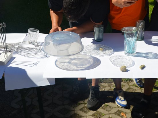 Personen führen im Freien ein Experiment durch. Auf einem weißen Tisch liegen verschiedene Glasgegenstände, ein Schneebesen und kleine Steine.