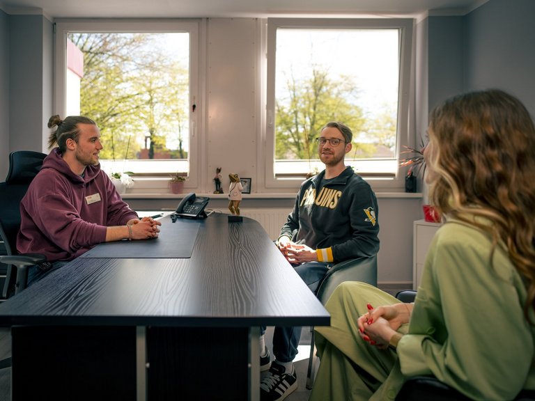 Drei Personen führen ein Gespräch in einem hellen Büro. Ein Mann im lila Hoodie sitzt hinter einem Schreibtisch. Ihm gegenüber sitzen ein Mann mit Brille in einem grauen Sweatshirt und eine Frau in grüner Kleidung. Tageslicht fällt durch die Fenster im Hintergrund.