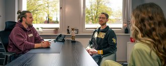 Drei Personen führen ein Gespräch in einem hellen Büro. Ein Mann im lila Hoodie sitzt hinter einem Schreibtisch. Ihm gegenüber sitzen ein Mann mit Brille in einem grauen Sweatshirt und eine Frau in grüner Kleidung. Tageslicht fällt durch die Fenster im Hintergrund.