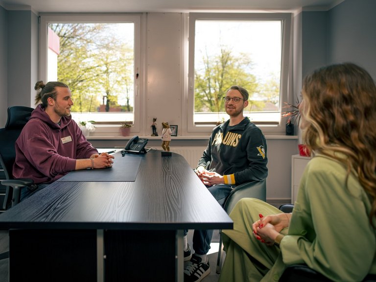 Drei Personen führen ein Gespräch in einem hellen Büro. Ein Mann im lila Hoodie sitzt hinter einem Schreibtisch. Ihm gegenüber sitzen ein Mann mit Brille in einem grauen Sweatshirt und eine Frau in grüner Kleidung. Tageslicht fällt durch die Fenster im Hintergrund.