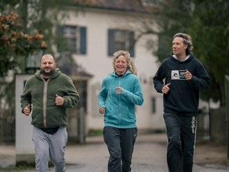 Drei lächelnde Personen joggen gemeinsam auf einer Straße. Links ein Mann in grüner Jacke, in der Mitte eine Frau in blauem Hoodie und rechts ein Mann in schwarzem Hoodie. Im Hintergrund ist ein weißes Haus zu sehen.