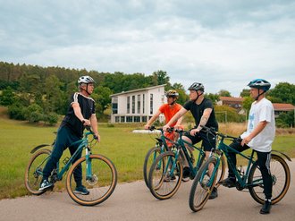 Vier junge Männer mit Fahrradhelm stehen mit ihren E-Bikes auf einem asphaltierten Weg und unterhalten sich. Im Hintergrund sind eine grüne Wiese, Bäume und ein weißes Gebäude zu sehen.