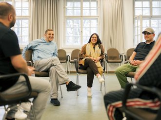 Eine Gruppe von Erwachsenen sitzt in einem hellen Raum mit großen Fenstern in einem Kreis auf Stühlen. Sie scheinen an einer Gruppendiskussion oder Therapiesitzung teilzunehmen.