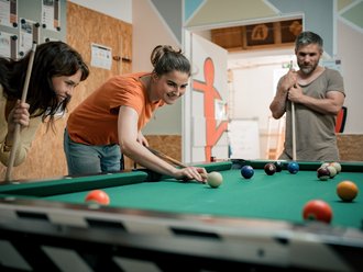 Zwei junge Frauen spielen Billard. Eine Frau im orangenen Shirt beugt sich zum Abstoß über den Tisch, während eine Freundin daneben steht und lächelnd zuschaut. Im Hintergrund beobachtet ein Mann das Spiel. Auf dem grünen Tuch liegen mehrere farbige Billardbälle.