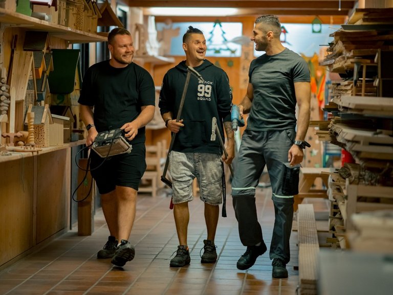 Drei Männer mit Werkzeugen in den Händen stehen in einer Werkstatt und unterhalten sich.
