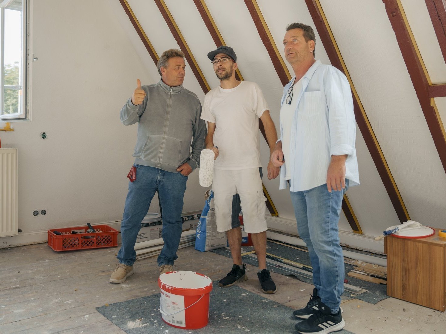 Drei Männer besprechen sich in einem Dachstuhl bezüglich des weiteren Vorgehens beim Anstrich der Wände.