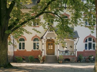 Ein weißes zweistöckiges Haus mit roten Ziegelakzenten und einem Holzeingang unter einer Veranda. Das Gebäude wird von großen Bäumen mit grünen Blättern gerahmt, und Topfpflanzen stehen an der Treppe.