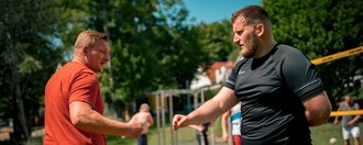 Zwei Männer stehen sich auf einem Sportplatz gegenüber. Der Mann im orangefarbenen T-Shirt und der Mann im schwarzen T-Shirt heben ihre Fäuste, als wollten sie sich die Fäuste stoßen. Im Hintergrund sind unscharf weitere Personen und Bäume zu erkennen.