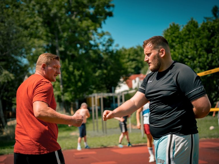 Zwei Männer stehen sich auf einem Sportplatz gegenüber. Der Mann im orangefarbenen T-Shirt und der Mann im schwarzen T-Shirt heben ihre Fäuste, als wollten sie sich die Fäuste stoßen. Im Hintergrund sind unscharf weitere Personen und Bäume zu erkennen.