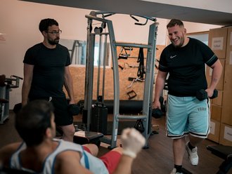 Drei Männer trainieren gemeinsam in einem Fitnessstudio. Ein Mann rechts lächelt breit und hält Hanteln, während er geht. Links steht ein weiterer Mann mit Brille und Hanteln. Im Vordergrund liegt ein dritter Mann auf einer Trainingsbank und führt eine Übung aus.