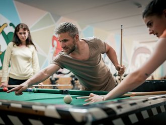 Ein Mann mit grauem Bart und eine Frau im Vordergrund spielen konzentriert Billard an einem grünen Tisch. Der Mann zielt auf den weißen Ball, während die Frau ihren Queue bereithält. Eine weitere Frau beobachtet das Spiel im Hintergrund in einem hellen Raum.