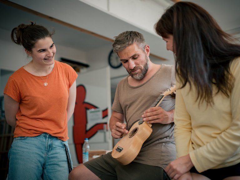 Drei Personen sitzen im Kreis. Ein Mann in der Mitte spielt Ukulele. Eine Frau links im orangen T-Shirt und eine Frau rechts im gelben Oberteil schauen ihm lächelnd zu.