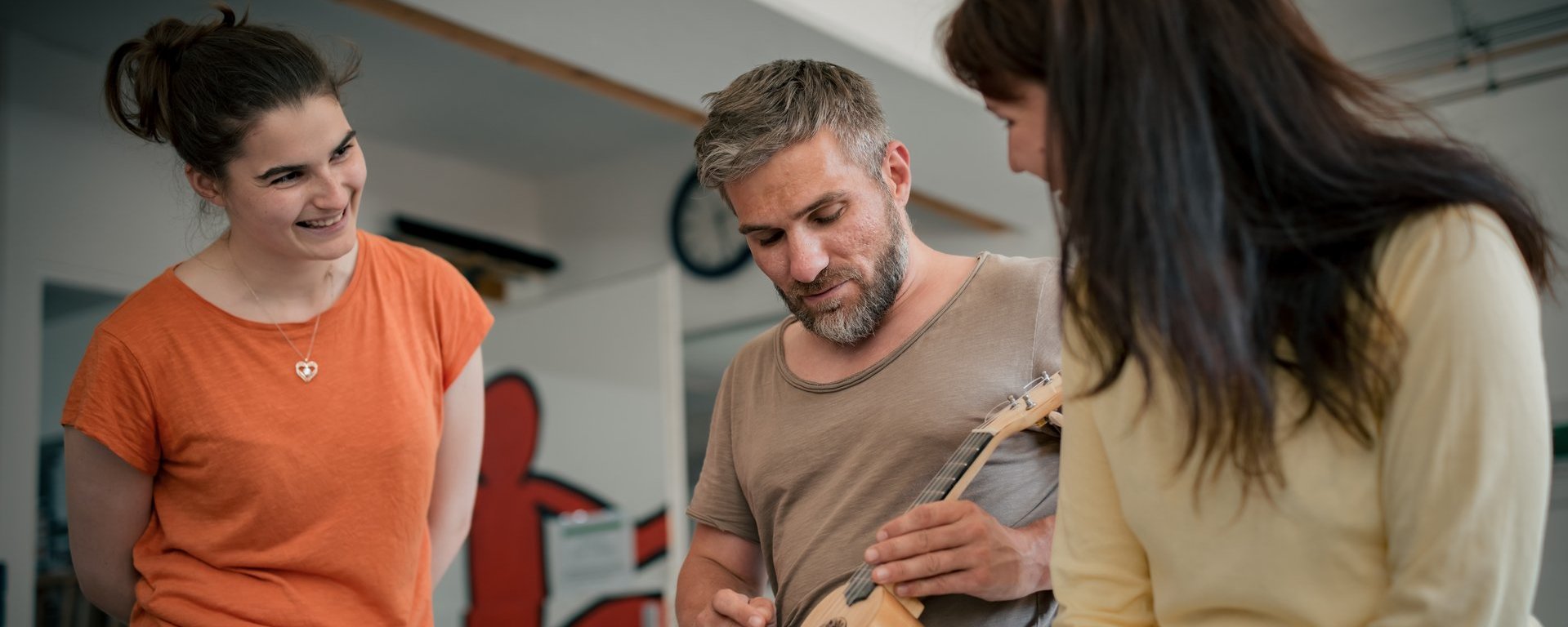Drei Personen sitzen im Kreis. Ein Mann in der Mitte spielt Ukulele. Eine Frau links im orangen T-Shirt und eine Frau rechts im gelben Oberteil schauen ihm lächelnd zu.