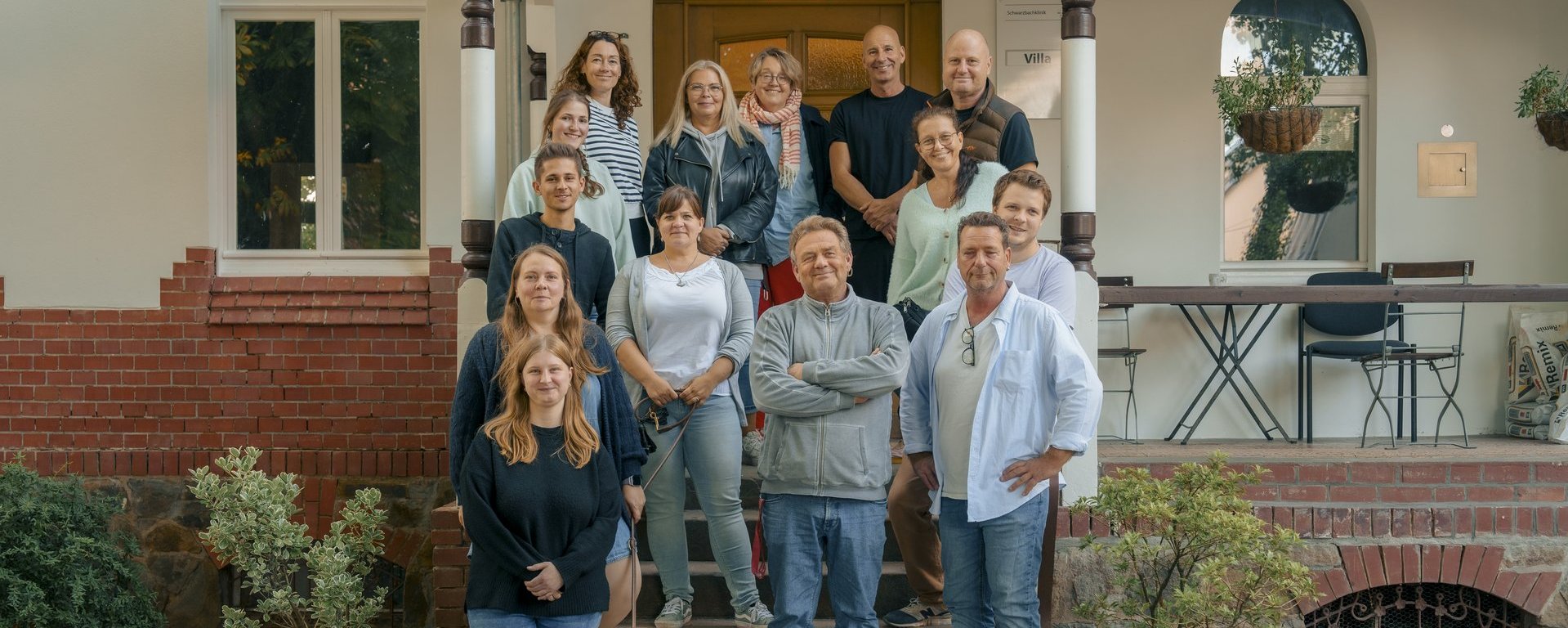 Eine Gruppe von etwa 15 Personen und ein Hund posieren lächelnd auf den Stufen vor einem hellen Gebäude. Die Menschen stehen in mehreren Reihen beieinander, der braune Hund sitzt im Vordergrund auf dem unteren Treppenabsatz.