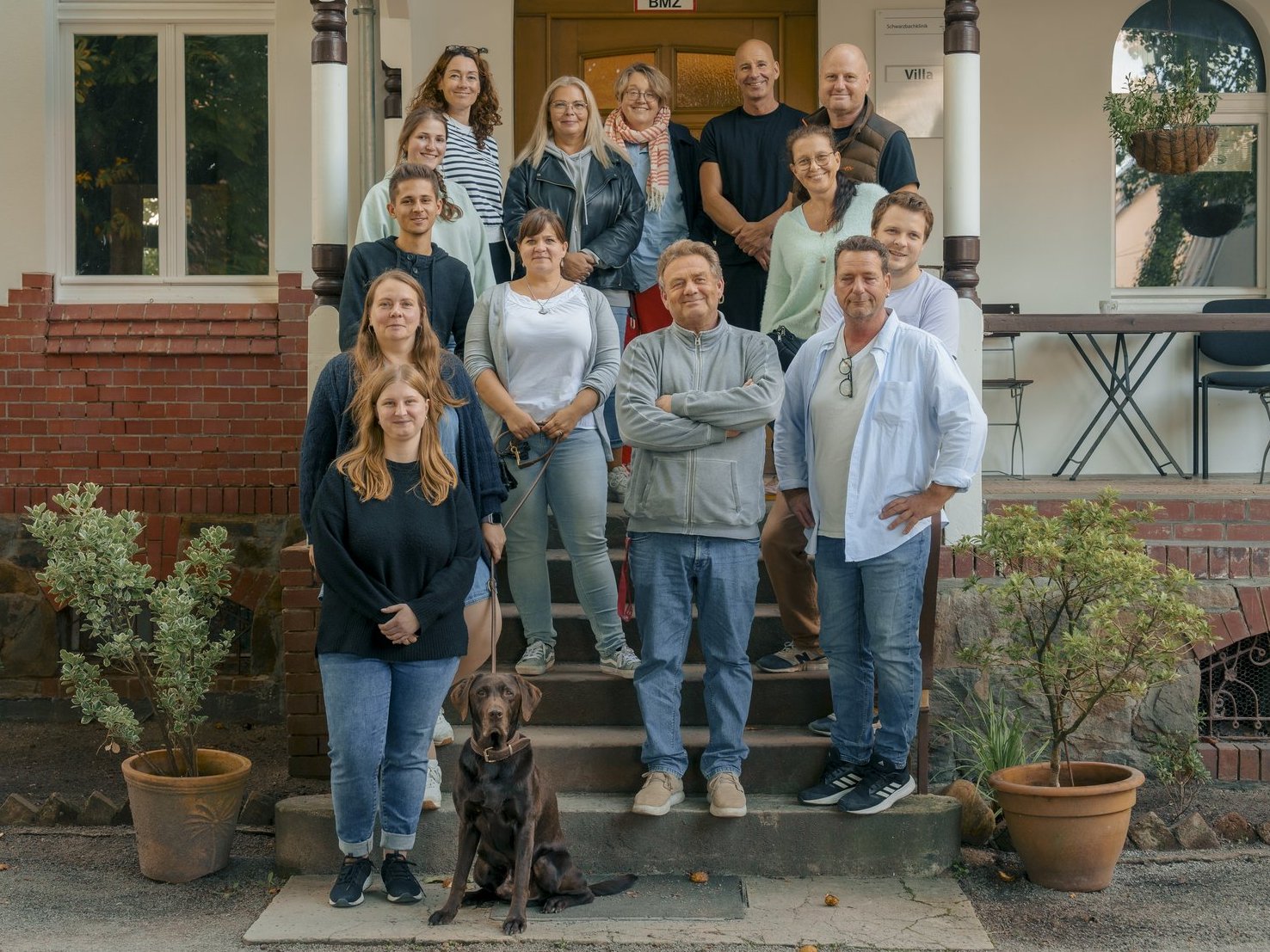 Eine Gruppe von etwa 15 Personen und ein Hund posieren lächelnd auf den Stufen vor einem hellen Gebäude. Die Menschen stehen in mehreren Reihen beieinander, der braune Hund sitzt im Vordergrund auf dem unteren Treppenabsatz.