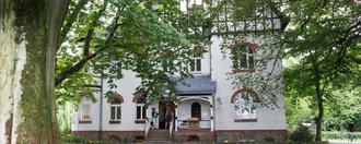 Aussenansicht: Gebäude mit spitzen Dächern und einem Eingang mit sechs Treppenstufen nach oben - im Verdergrund sehr alte große Bäume