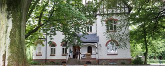 Schwarzbachklinik Aussenansicht: Gebäude mit spitzen Dächern und einem Eingang mit sechs Treppenstufen nach oben - im Verdergrund sehr alte große Bäume