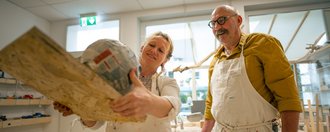 Zwei Künstler in schmutzigen weißen Kitteln stehen in einer Werkstatt. Die Frau hält ein großes Kunstobjekt aus Holz und Papiermache hoch und lächelt, während der Mann mit Brille und Schnurrbart ihr zufrieden zuschaut.