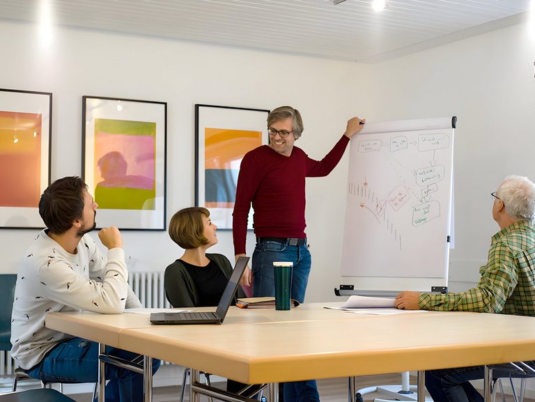 Besprechungsraum: ein junger Mann steht an einem Flipchart und erklärt einer kleinen Gruppe lächelnd sein "Gekritzel"