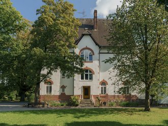 Ein weißes, zweistöckiges Haus mit einem dunklen Satteldach und roten Ziegelsteinen als Akzent an den Fenstern. Das Gebäude steht zentral auf einer grünen Wiese, umgeben von hohen Laubbäumen unter einem blauen Himmel.