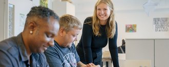 Drei Personen arbeiten gemeinsam an einem Tisch. Zwei Männer sitzen und knüpfen ein Armband, während eine Frau lächelnd neben einem Tisch steht. Die Szene zeigt eine freundliche, kooperative Zusammenarbeit in einem hellen Raum.
