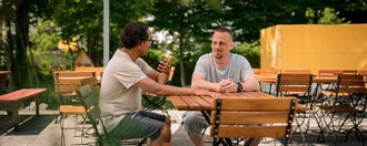 Zwei Männer sitzen an einem Holztisch in einem Außenbereich und unterhalten sich. Der Mann links hält eine Flasche in der Hand, während der Mann rechts ihm zuhört.