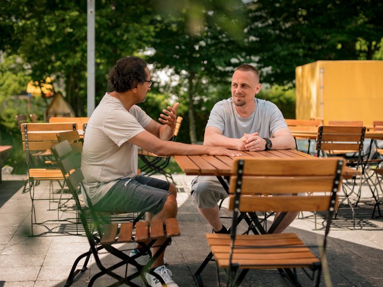 Zwei Männer sitzen an einem Holztisch in einem Außenbereich und unterhalten sich. Der Mann links hält eine Flasche in der Hand, während der Mann rechts ihm zuhört.