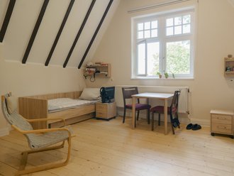 Ein helles Zimmer mit Schrägdach und dunklen Holzbalken. Der Raum hat einen hellen Holzboden, zwei Einzelbetten, einen Schreibtisch mit zwei Stühlen und einen Schaukelstuhl. Ein großes Fenster lässt Tageslicht herein.