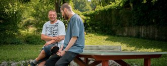 Zwei Männer sitzen lächelnd auf einem roten Tischtennistisch im Freien und unterhalten sich. Sie befinden sich in einer grünen Parkanlage mit Bäumen im Hintergrund.
