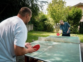 Zwei Männer spielen Tischtennis im Garten. Der Mann im Vordergrund trägt ein weißes T-Shirt, sein Gegenspieler gegenüber ein blaues. Beide halten rote Schläger und konzentrieren sich auf das Spiel am grünen Tischtennistisch.