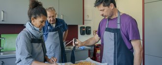 Drei Menschen in Schürzen bereiten in einer Küche gemeinsam Essen zu. Ein Mann hält einen Spachtel über ein Backblech mit einem gebackenen Gericht, während eine Frau und ein älterer Mann danebenstehen und zuschauen.